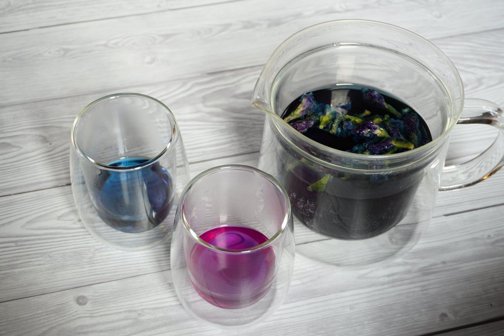 Teekanne mit Blüten in dunkelblau gefärbtem Tee, ein Teeglas mit blauer Flüssigkeit, eines mit purpur-violetter Flüssigkeit